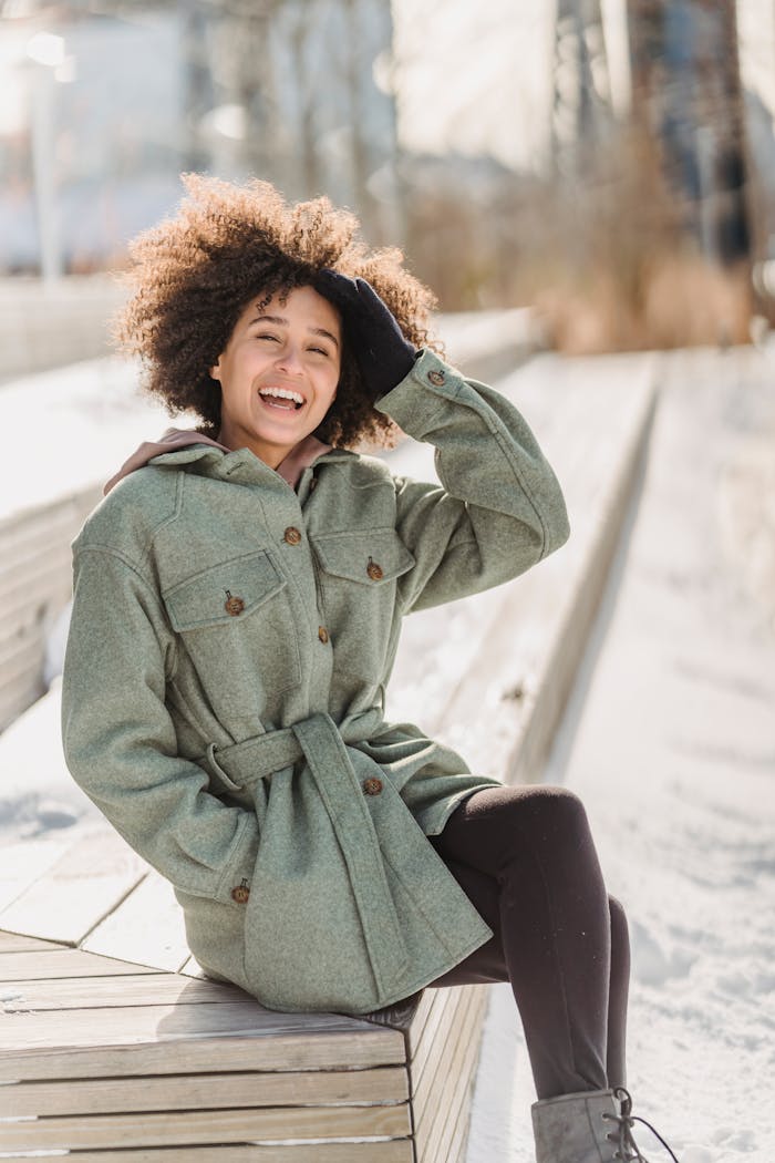 Happy young ethnic female wearing warm coat touching curly hair and looking at camera while sitting on wooden bench in snowy park on cold winter day